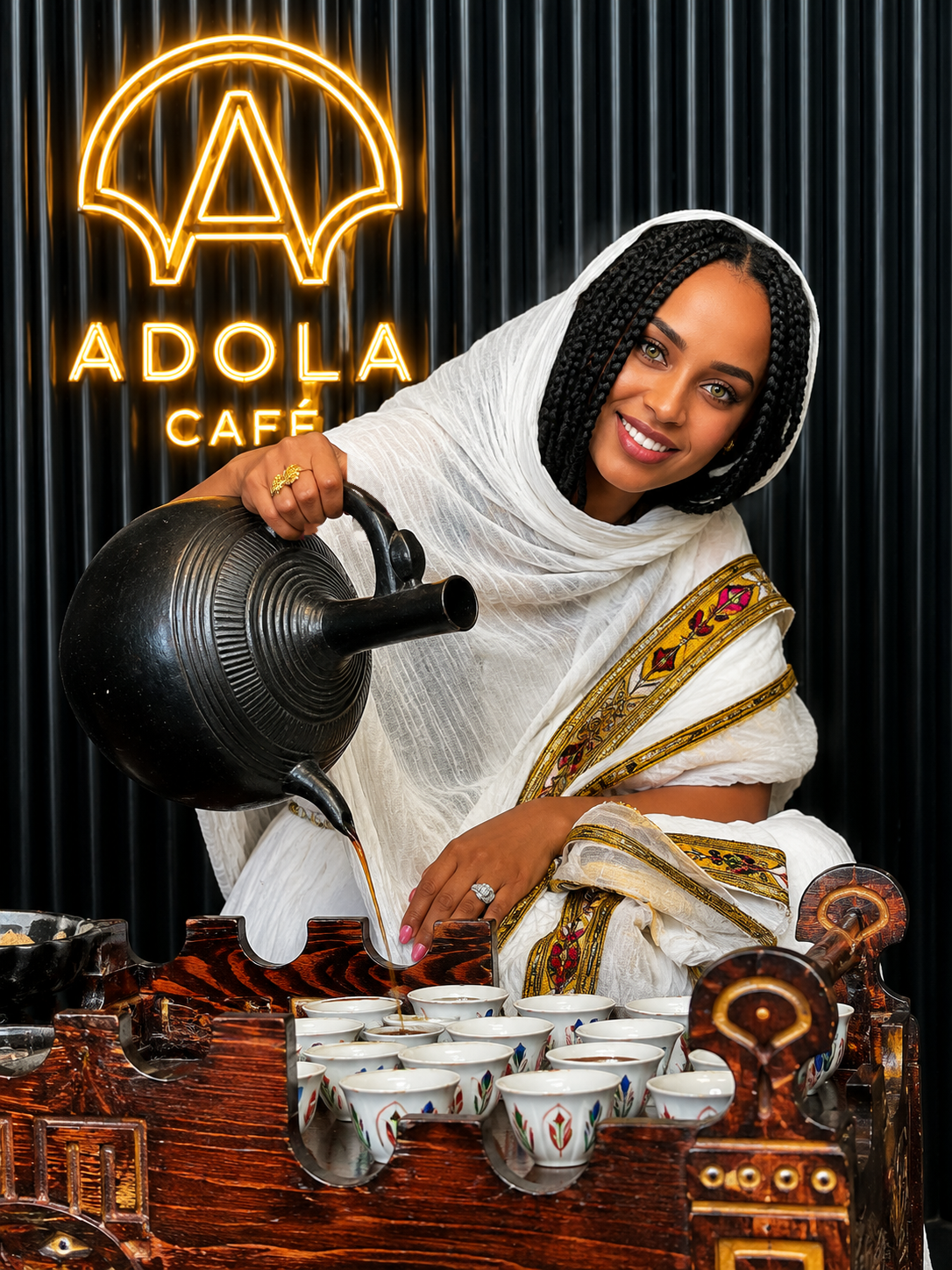 A woman in traditional Ethiopian dress pouring coffee from a jebena beneath the ADOLA Café neon sign